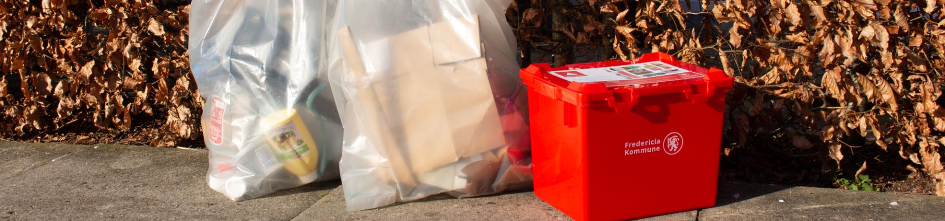 Rød kasse til farligt affald foran hæk sammen med sække til genbrug. 